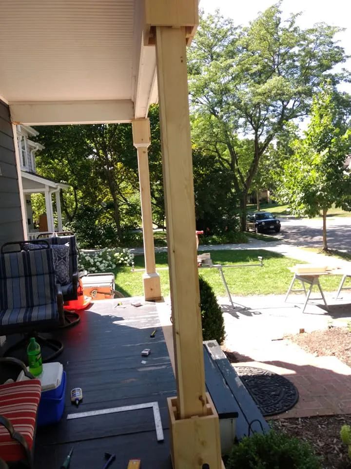 Porch columns under construction on a dark porch; a green yard and street are in the background.