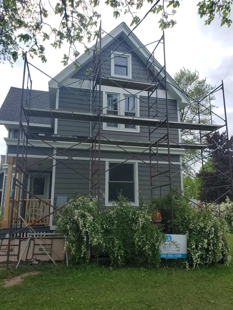 Gray house with scaffolding, undergoing renovations. White trim, bushes in front.