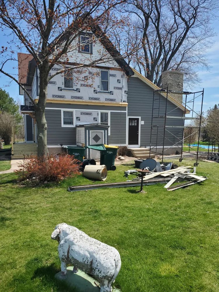 House exterior under renovation with new gray siding, scaffolding, and yard debris. A sheep statue is in the foreground.