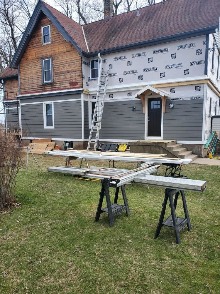 House exterior during renovation. Siding partially installed; ladder, sawhorses, and materials on the lawn.