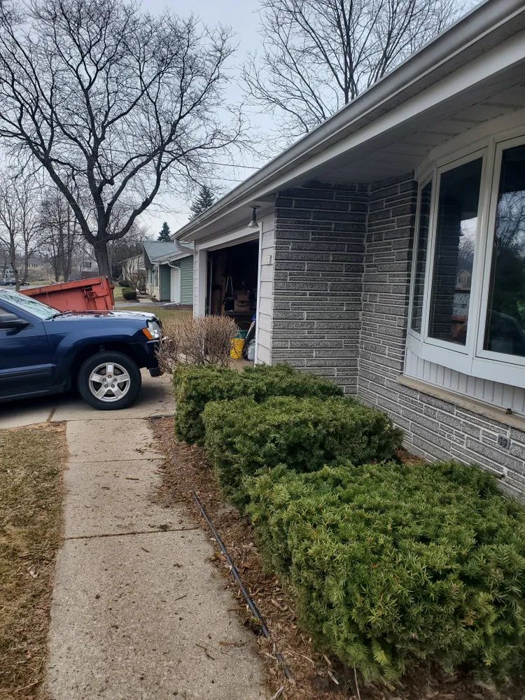Side view of a house with a garage and green bushes. A blue truck is parked nearby.