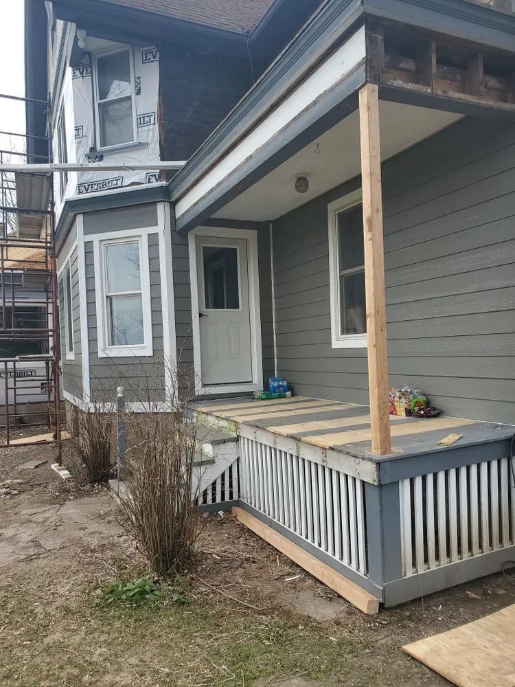 House exterior with gray siding, porch with railing, door, and unfinished construction.
