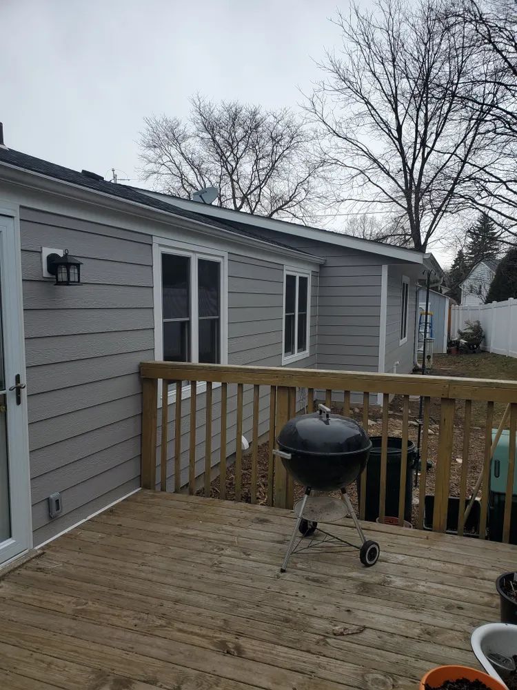 Gray house with a wooden deck, a black grill, and bare trees in the background.