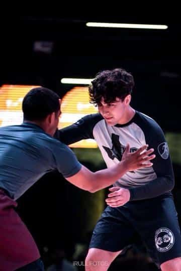 Two wrestlers in a mat area, one in a black-and-white shirt grappling with another in a blue top and maroon shorts.
