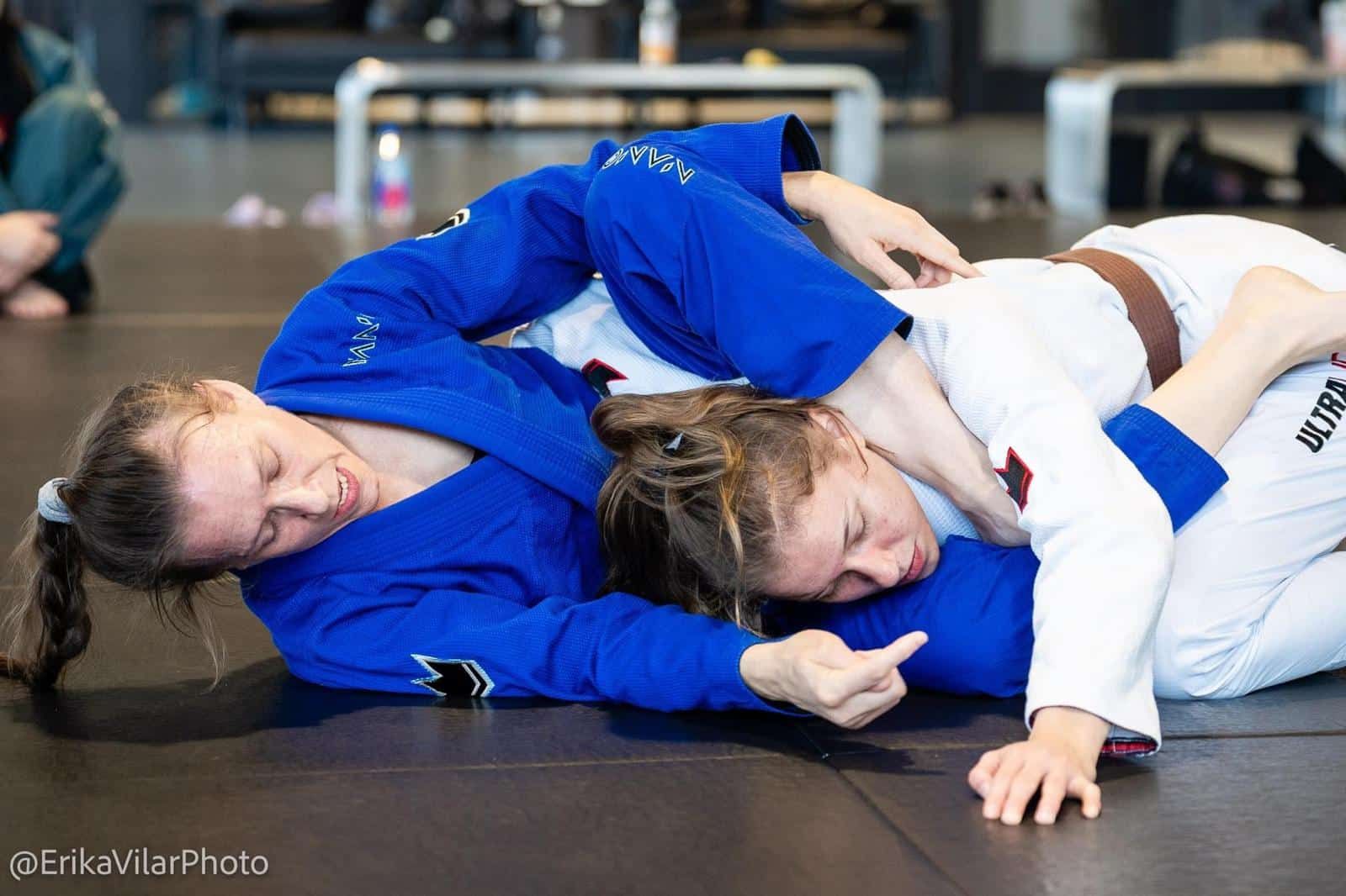Two judo grapplers in blue and white gis on a mat, one pinning the other during a ground hold.