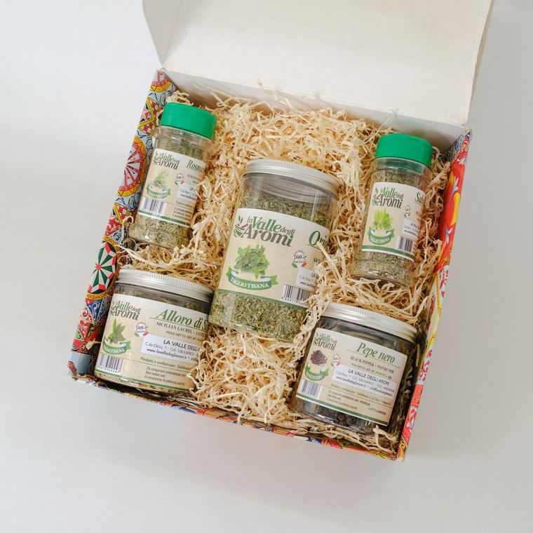Confezione regalo aperta con tre vasetti e due bottigliette di conserve su un letto di carta sminuzzata.