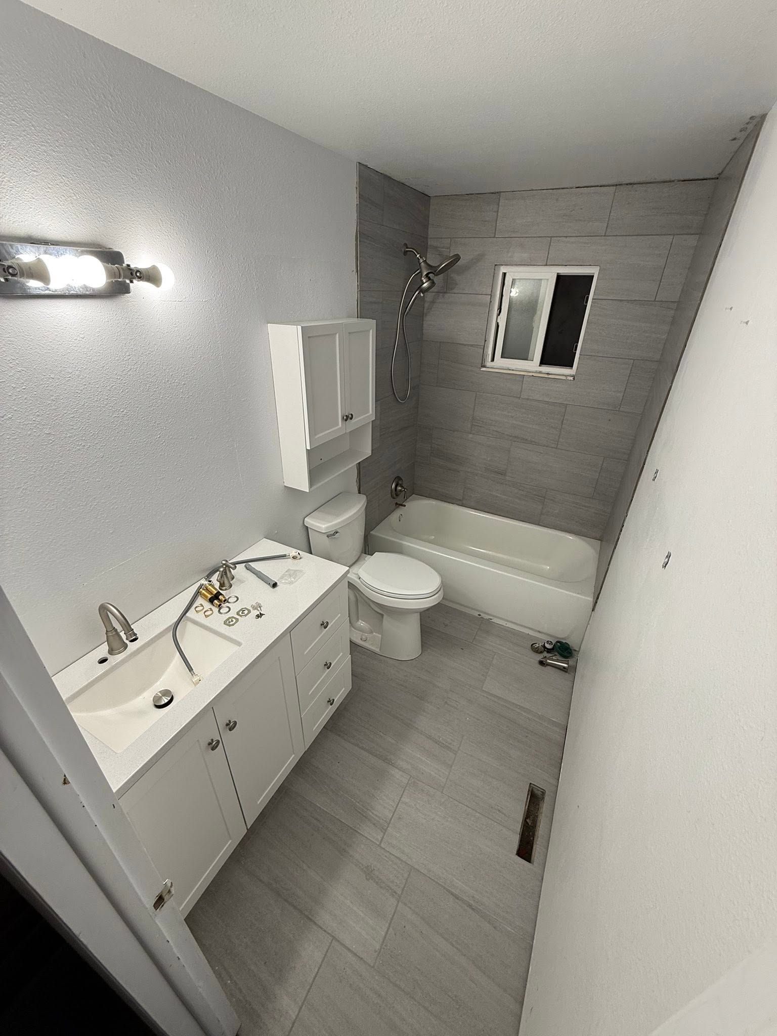 Narrow bathroom with white vanity, toilet, tub, gray tile walls, and wall mirror.