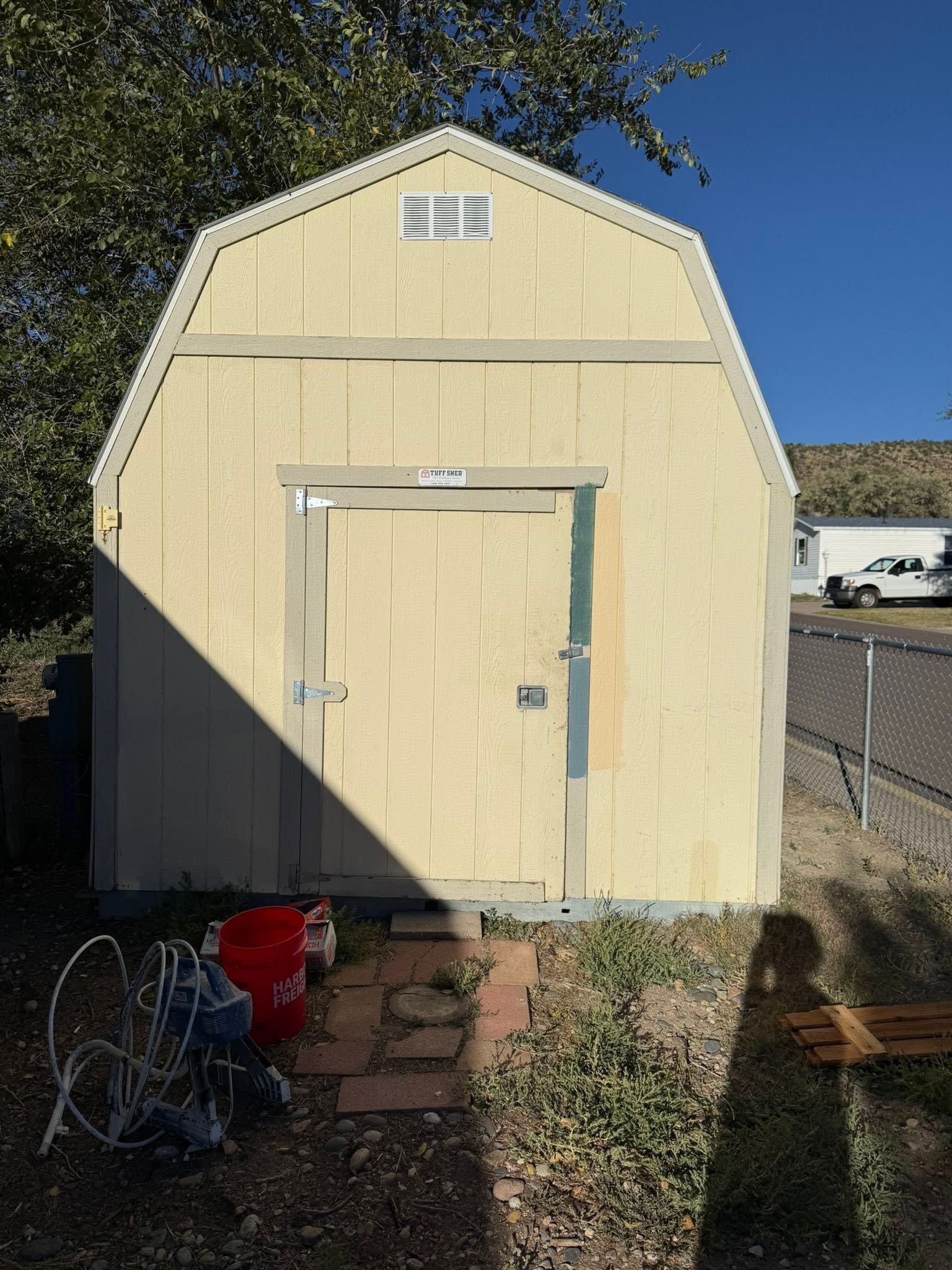 Small beige shed with a white door in a sunny backyard, with bikes and a red bucket in front.