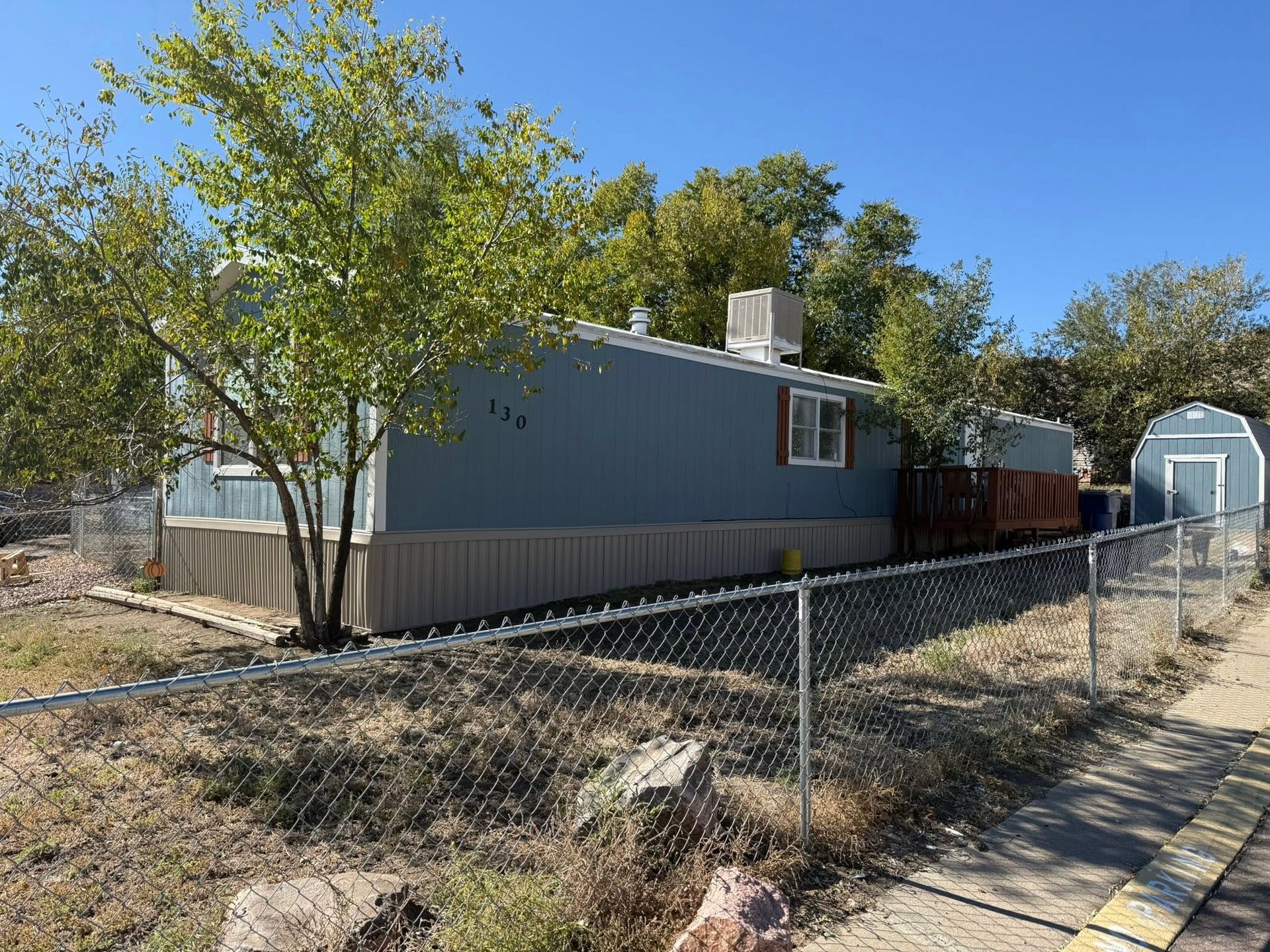 Blue mobile home behind a chain-link fence on a sunny day, with trees and a shed in the background