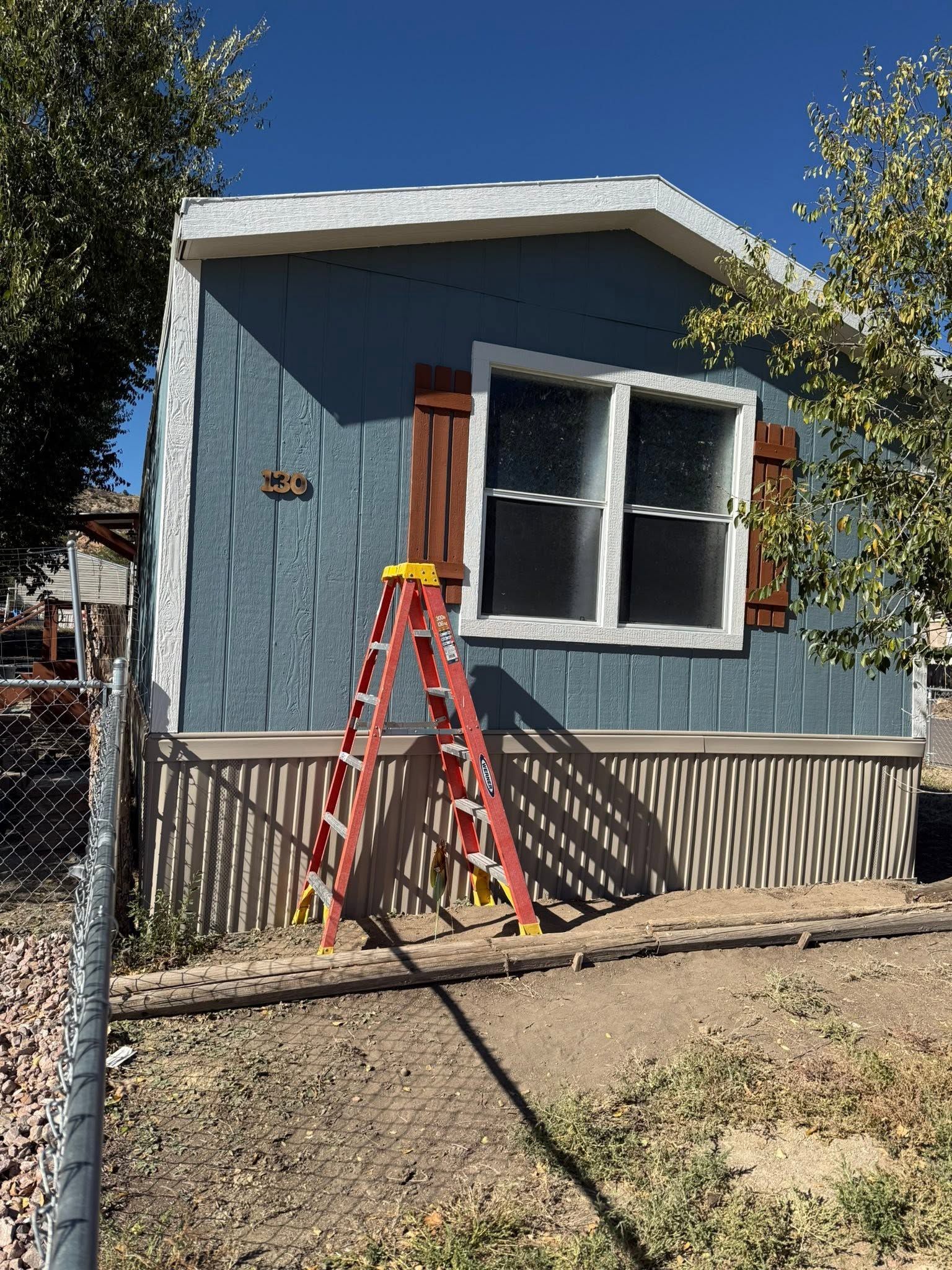Small blue house with white trim, a red ladder by the front window, and a fenced yard in sunlight