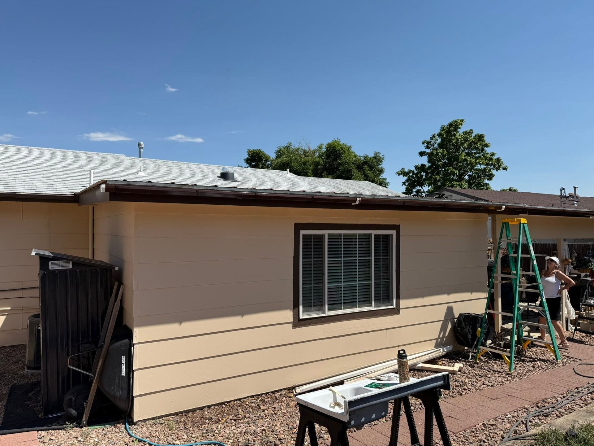Tan house exterior with workers on a ladder and tools in a gravel yard under a blue sky