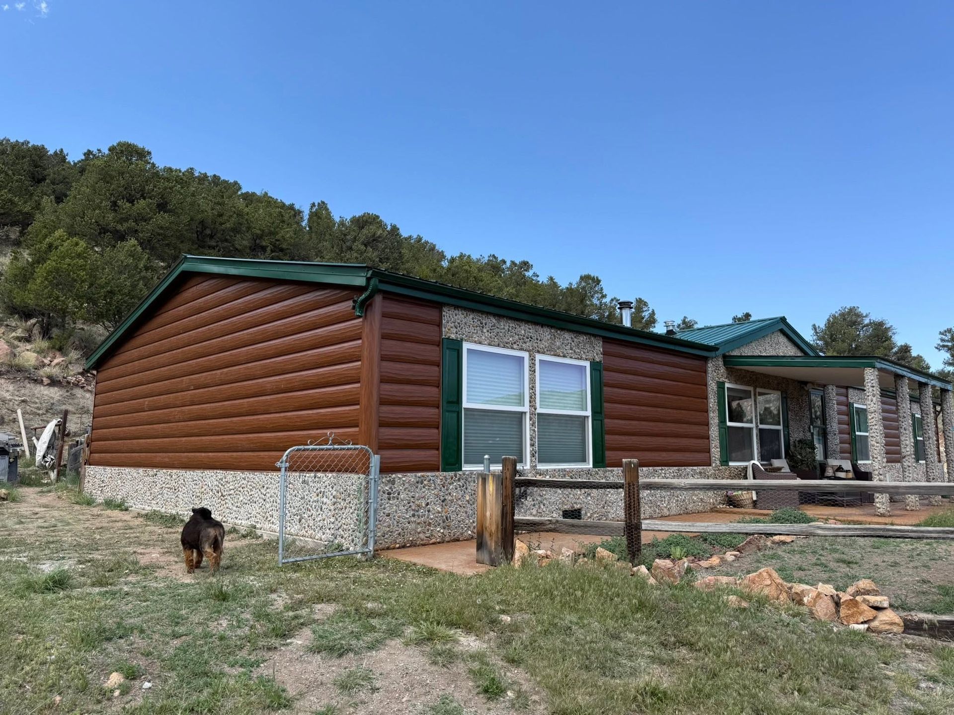Log cabin with green trim under a blue sky, with a small dog near a fenced yard.
