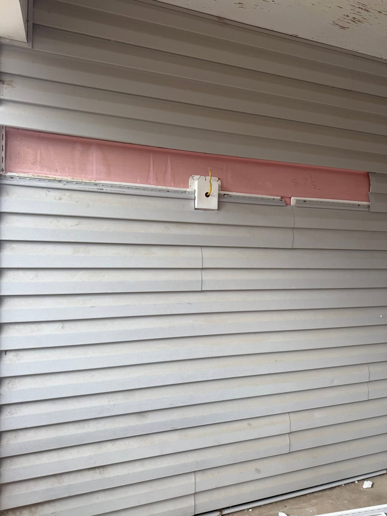 Closed gray siding with a pink band and small wall-mounted fixture under a porch roof