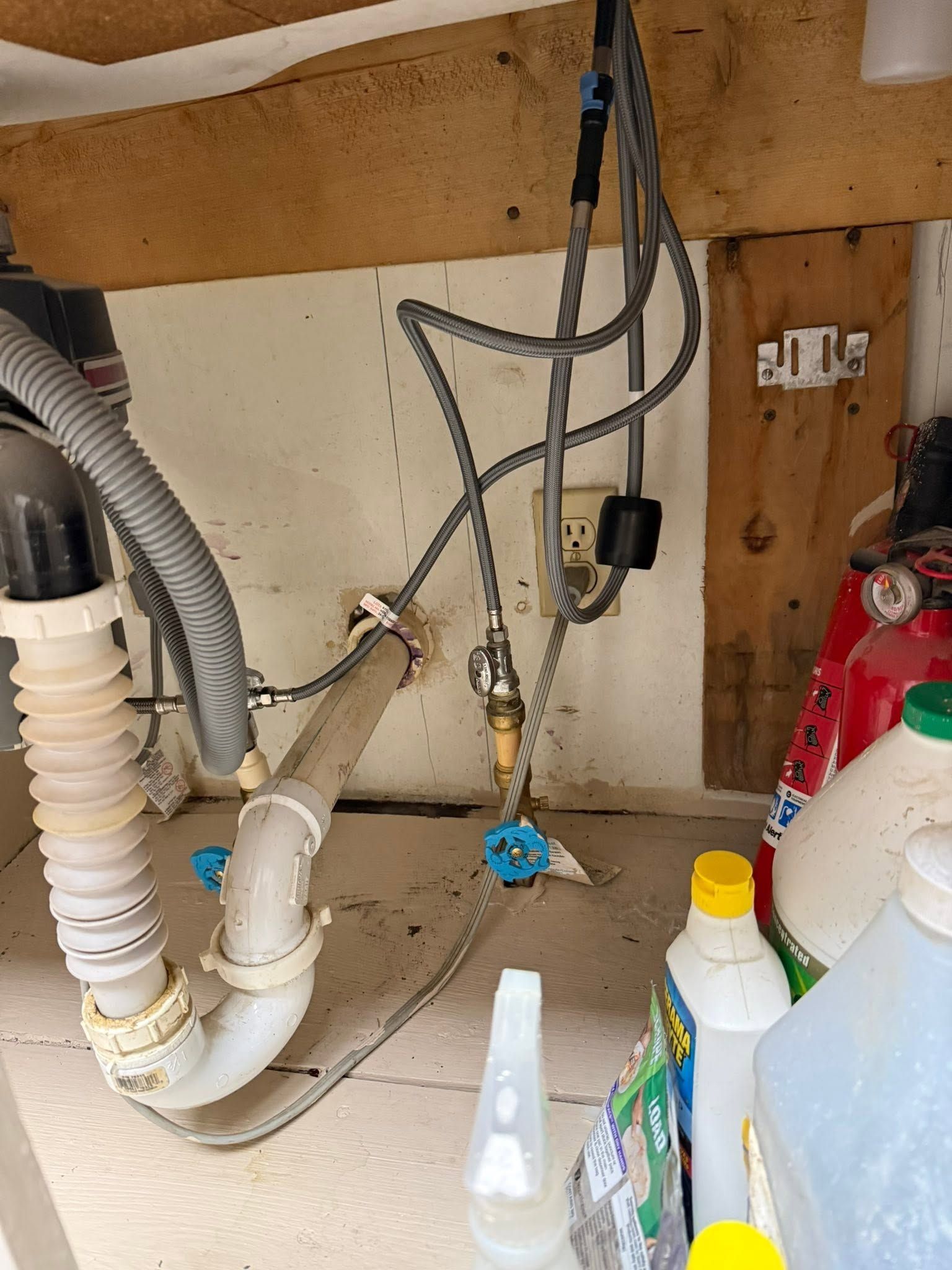 Under-sink plumbing with pipes, hoses, valves, and cleaning bottles in a cabinet with a leak sensor nearby