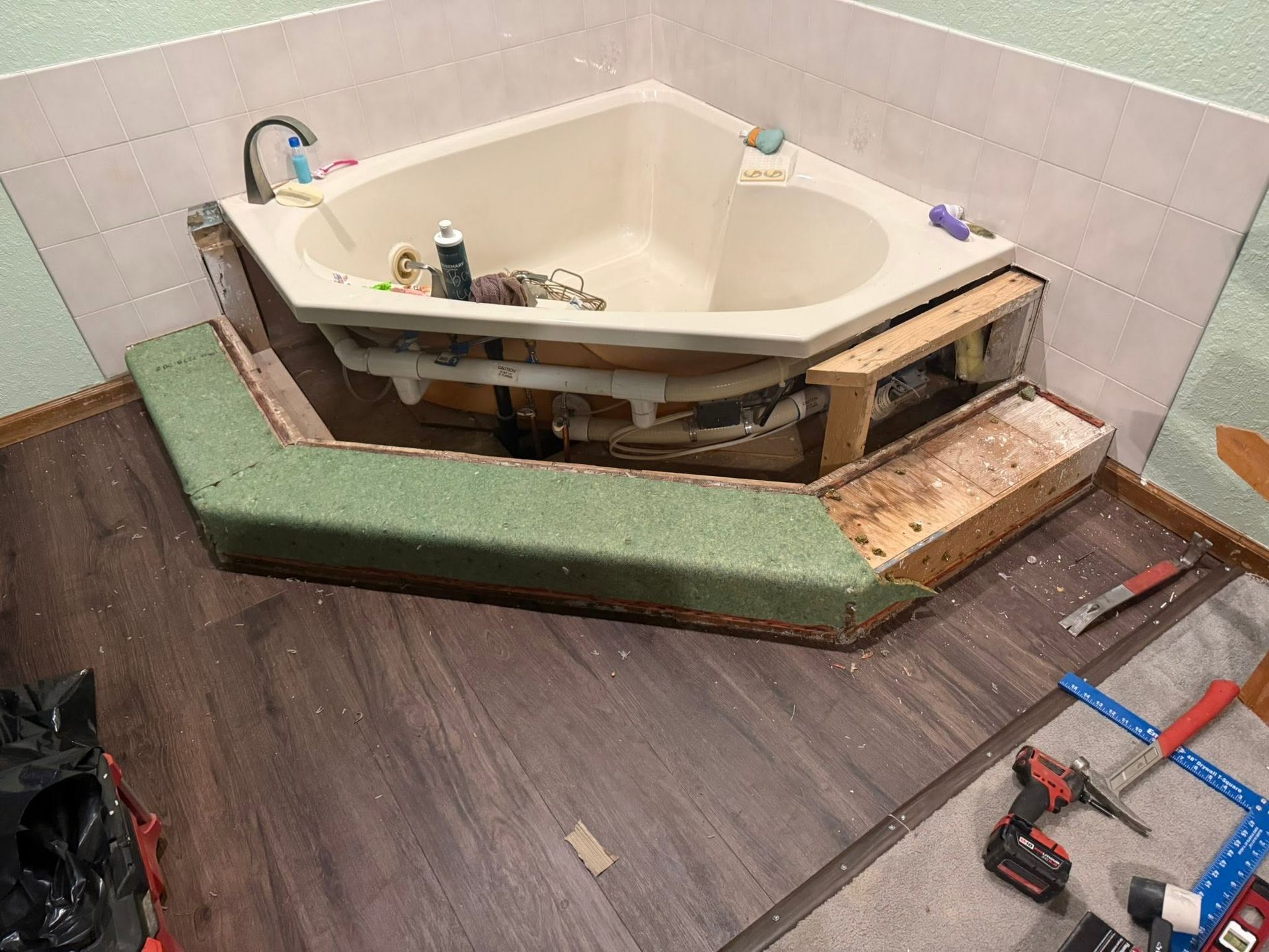 Damaged built-in bathtub under renovation, with exposed framing and tools on the bathroom floor.