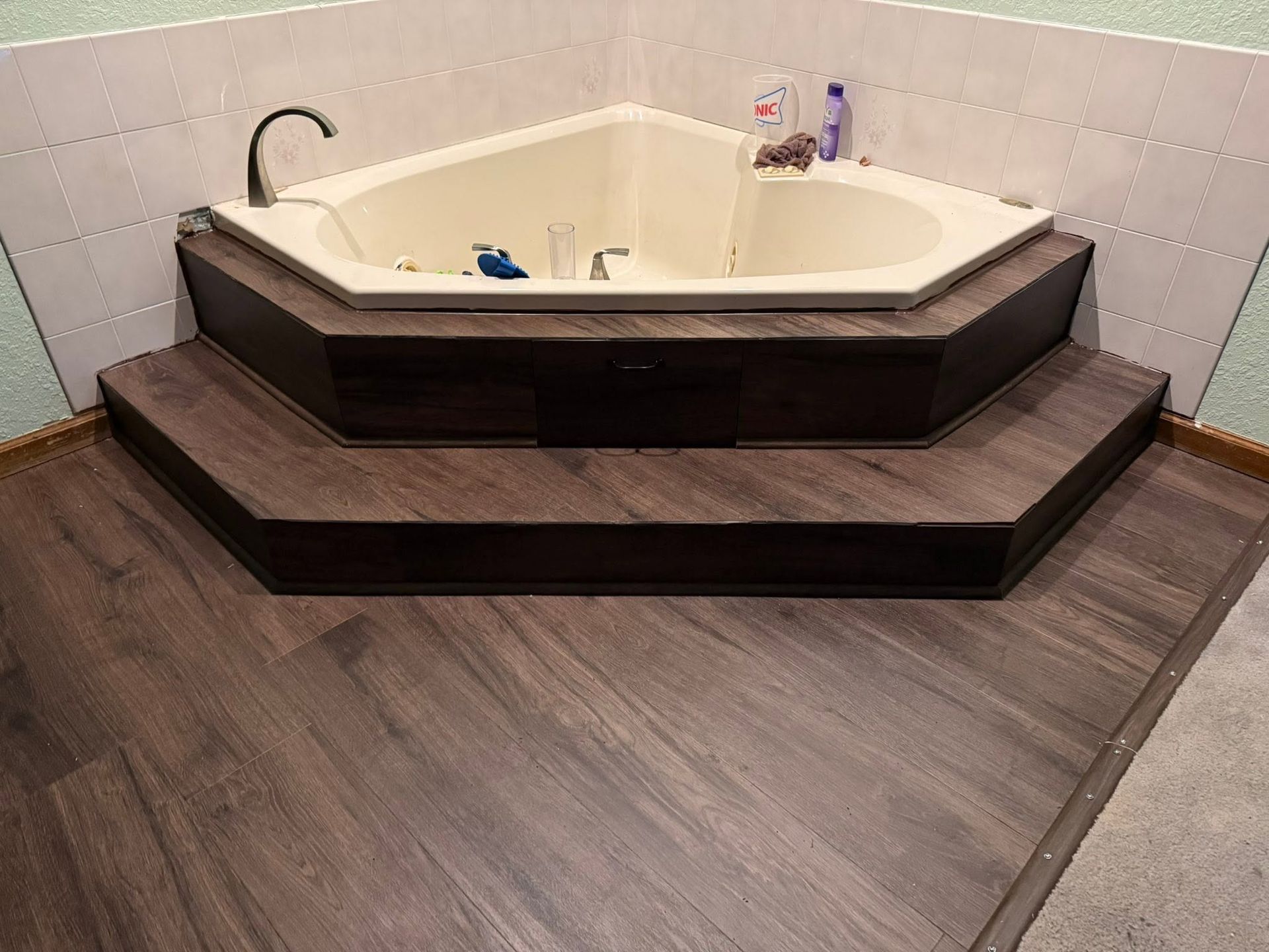 Octagonal indoor hot tub with beige tile and wood-look steps in a tiled room