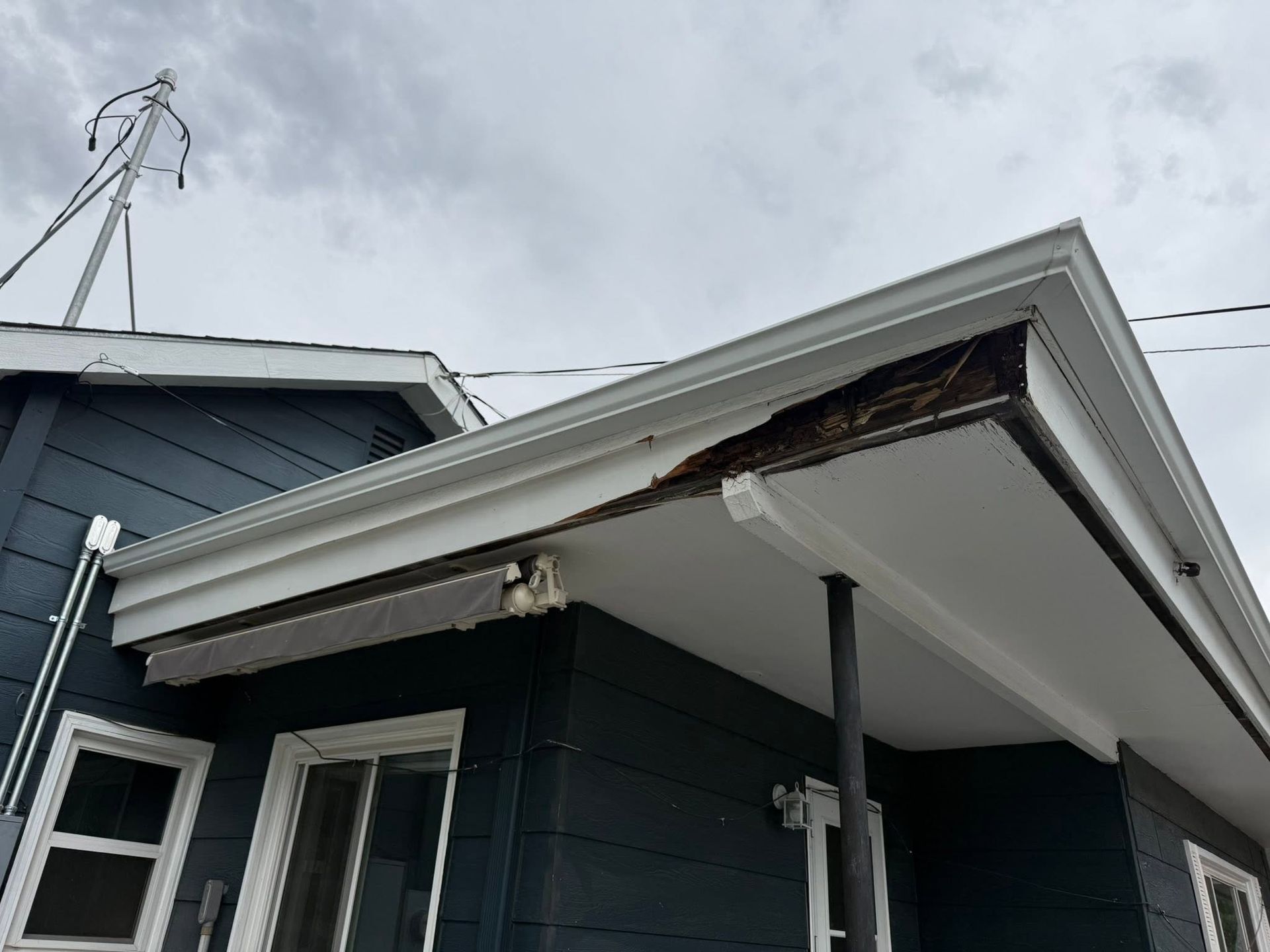 Damaged roof overhang on a dark blue house with exposed wood and a tilted gutter.