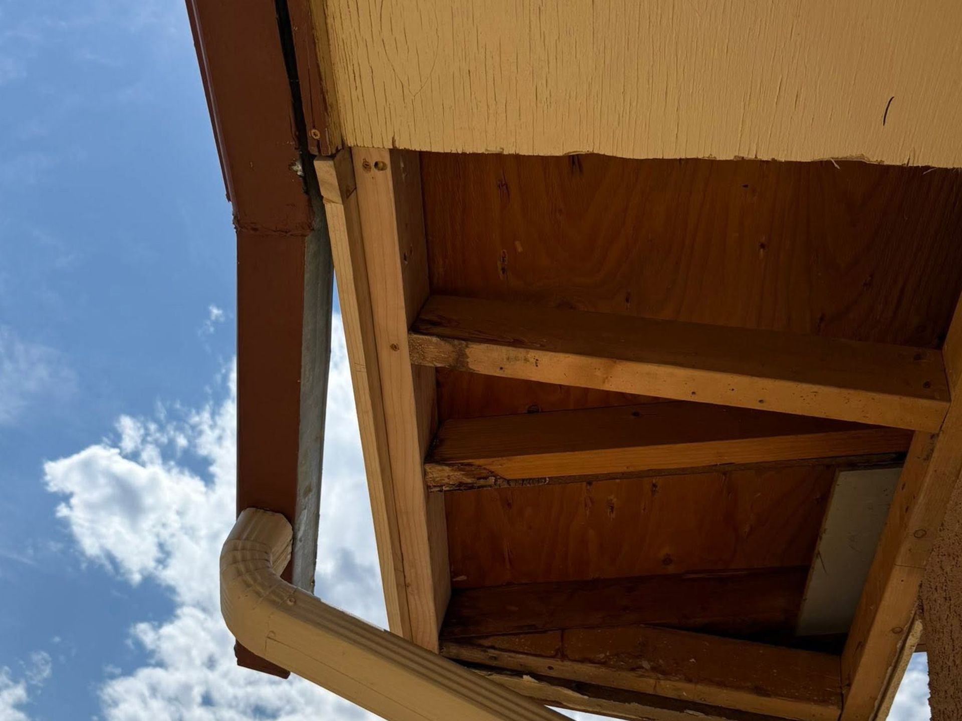Wooden roof overhang and support beams beneath a house, with blue sky and a small cloud visible.