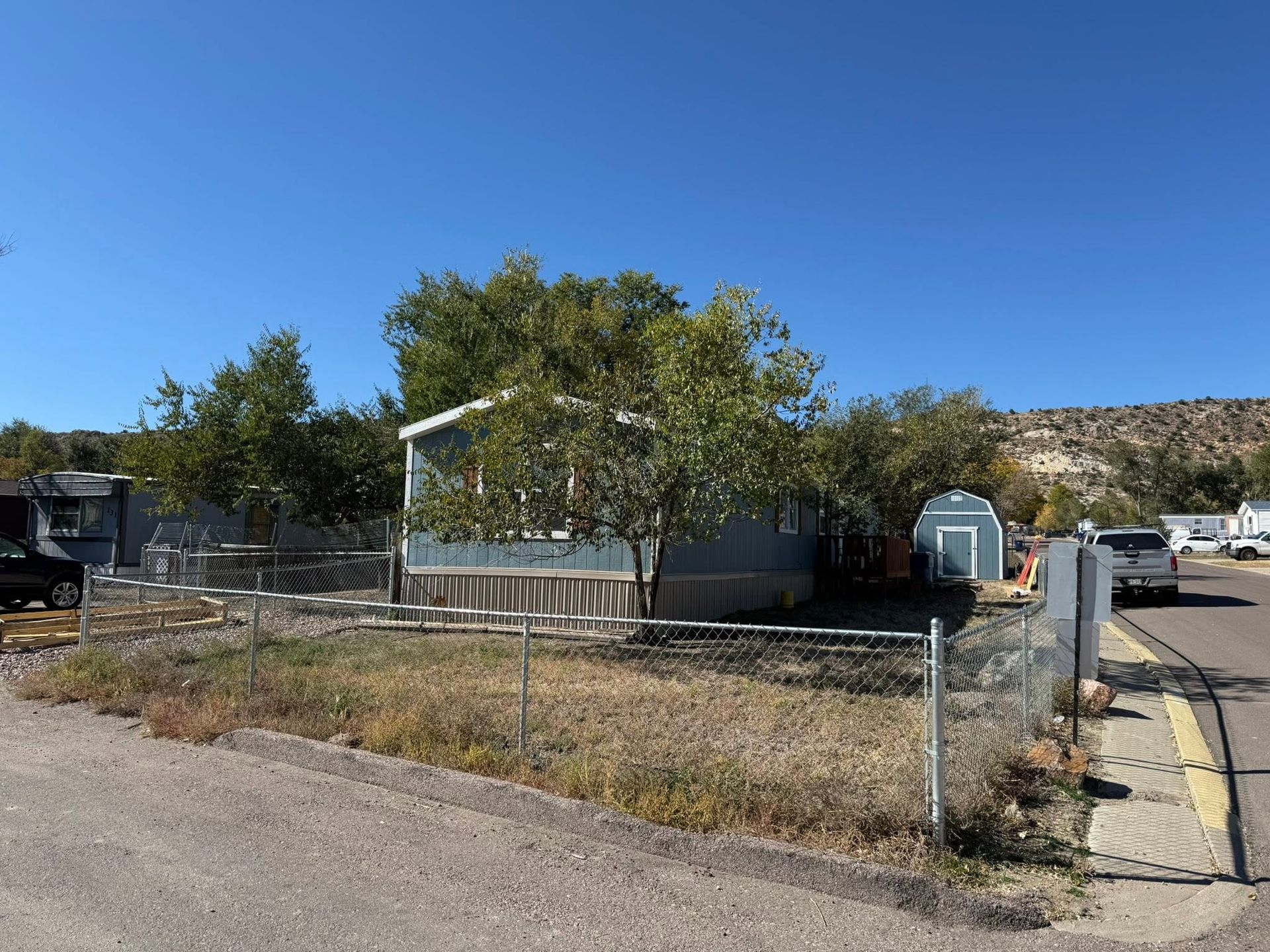 Empty fenced lot with a tree, chain-link fence, and small trailer beside a road under a blue sky