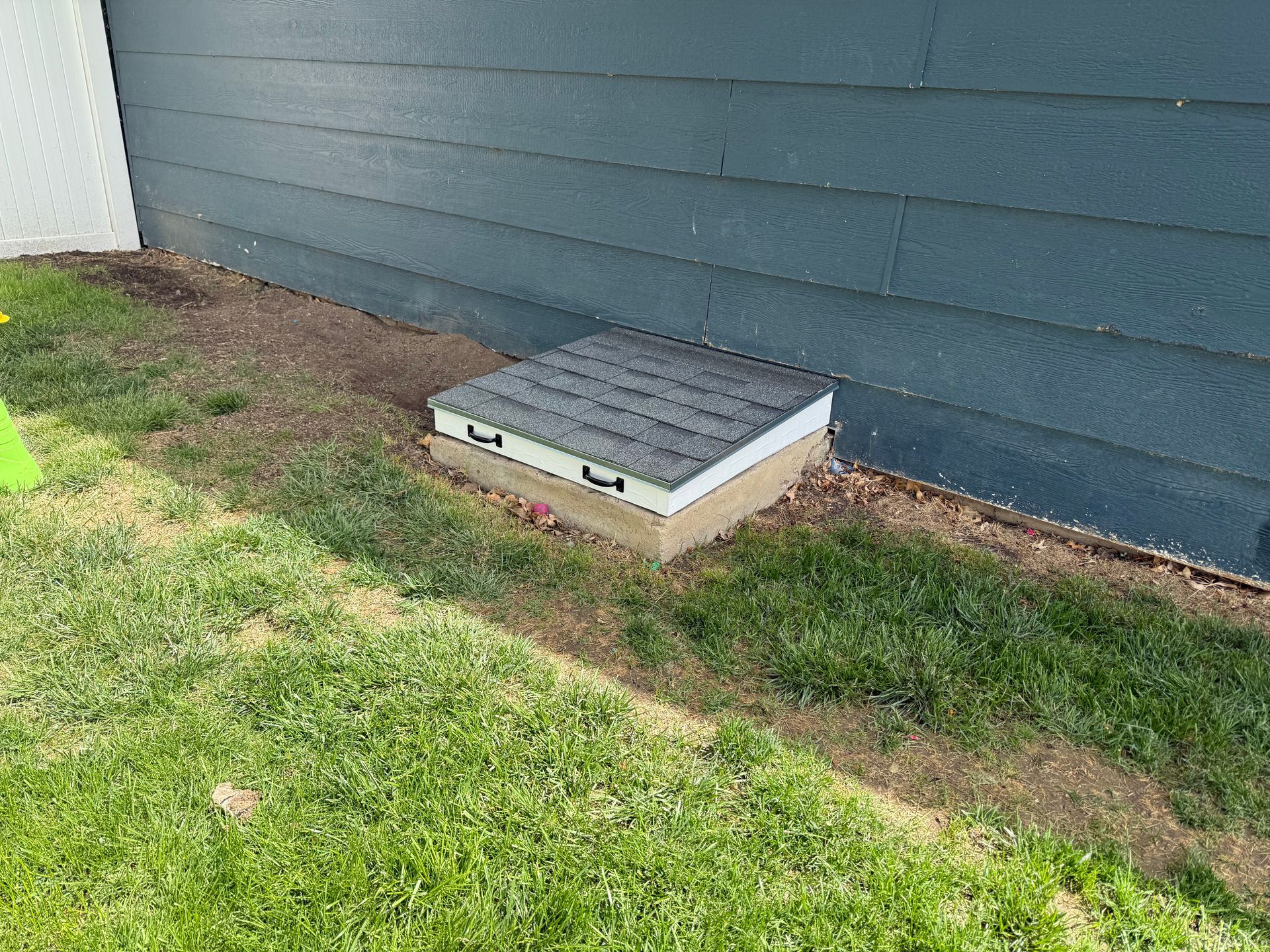 Small utility hatch with a gray cover beside a blue building on grass and dirt.