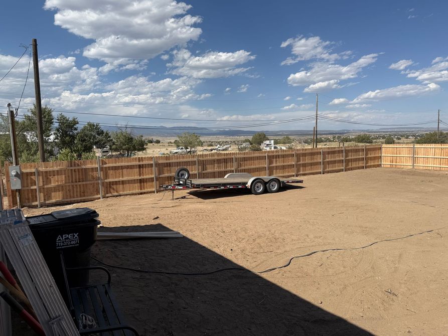 Open dirt lot with wooden fencing, a parked trailer, and cloudy sky in the distance