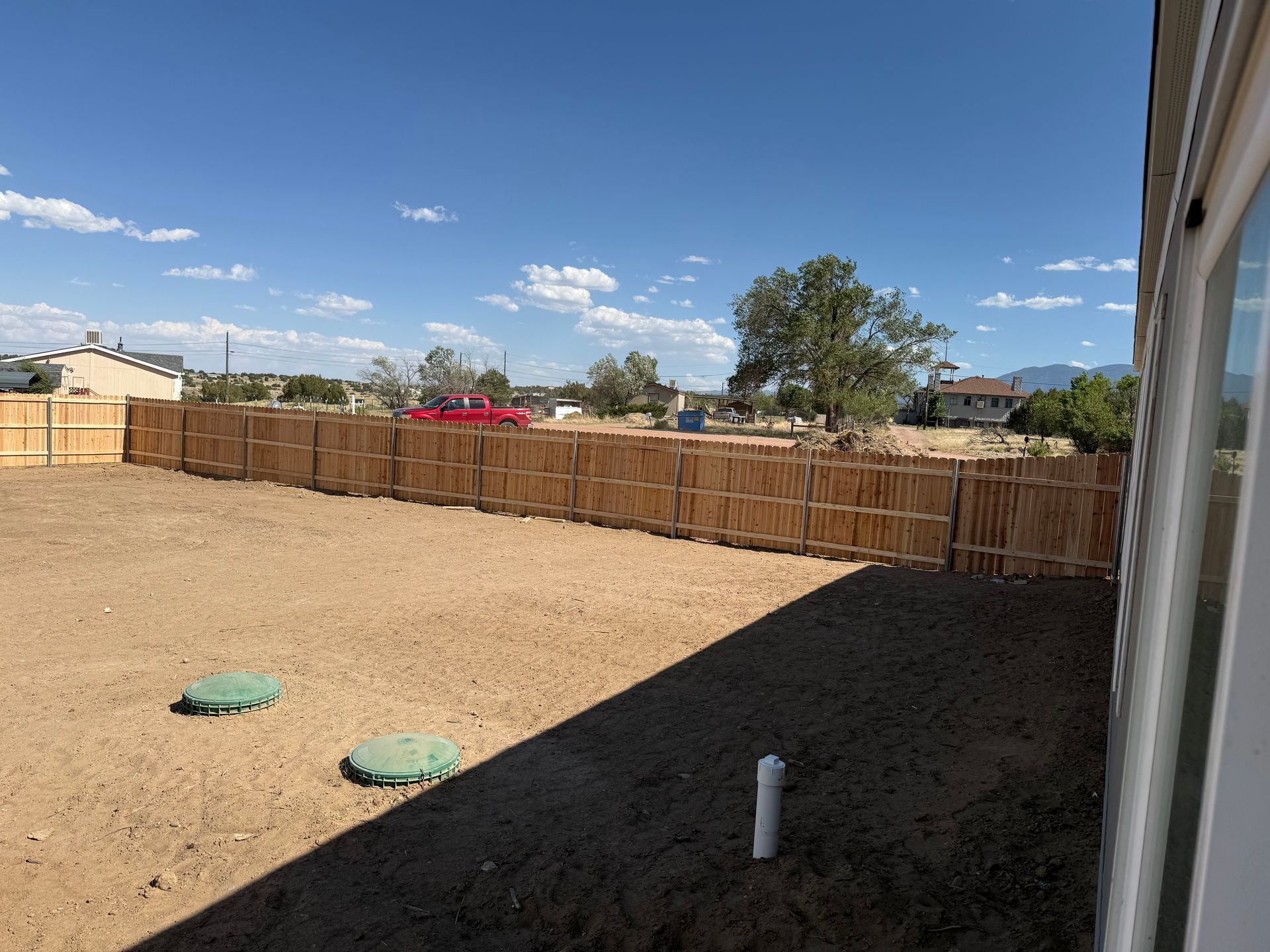 Dry backyard with gravel, wooden fence, green utility covers, and a shaded house wall under a blue sky.