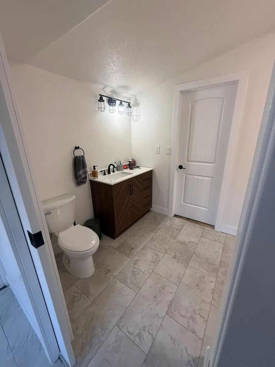 Small bathroom with toilet, wood vanity, mirror light, and gray tile floor.