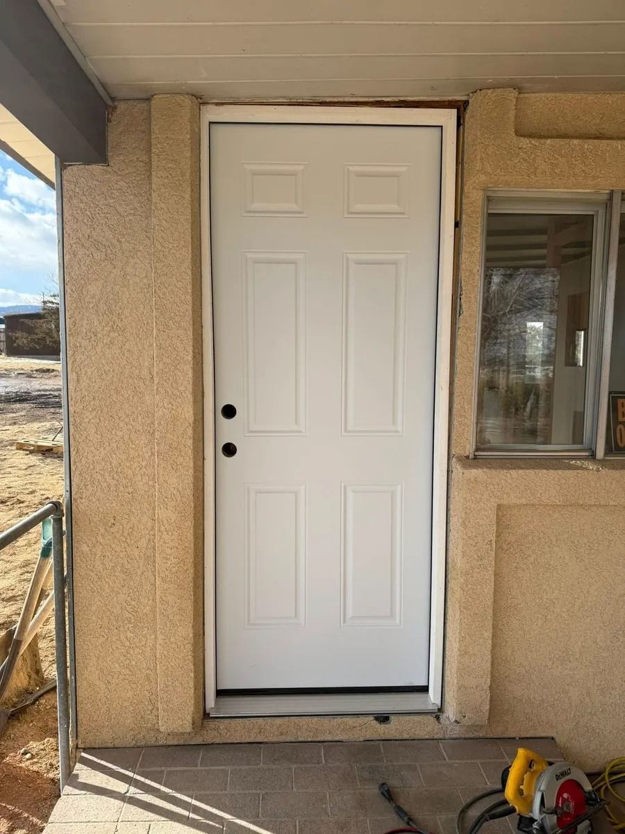 White front door with black knob on a tan stucco house porch, next to a window and construction tools.
