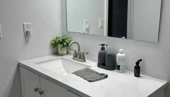 Modern bathroom vanity with white sink, gray countertop, mirror, soap dispensers, and a small plant.
