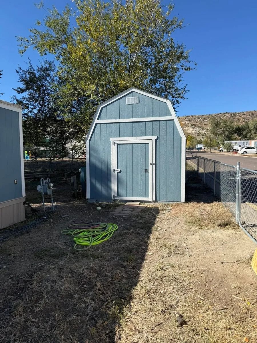 Blue garden shed in a fenced backyard with a tree and dry grass under a clear sky