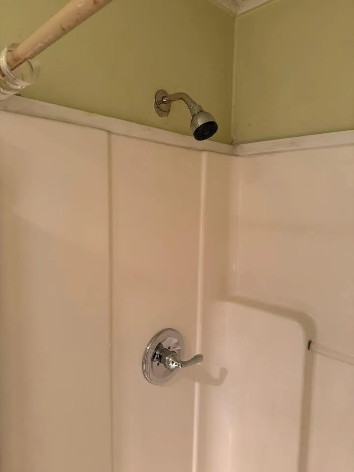 Empty beige shower stall with a wall-mounted showerhead and curtain rod visible near the ceiling