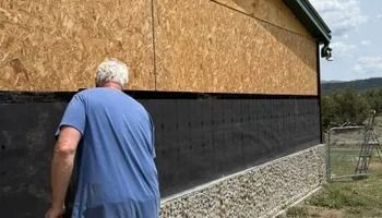 Man in blue shirt walking beside a house with wood siding and a black foundation on a sunny day