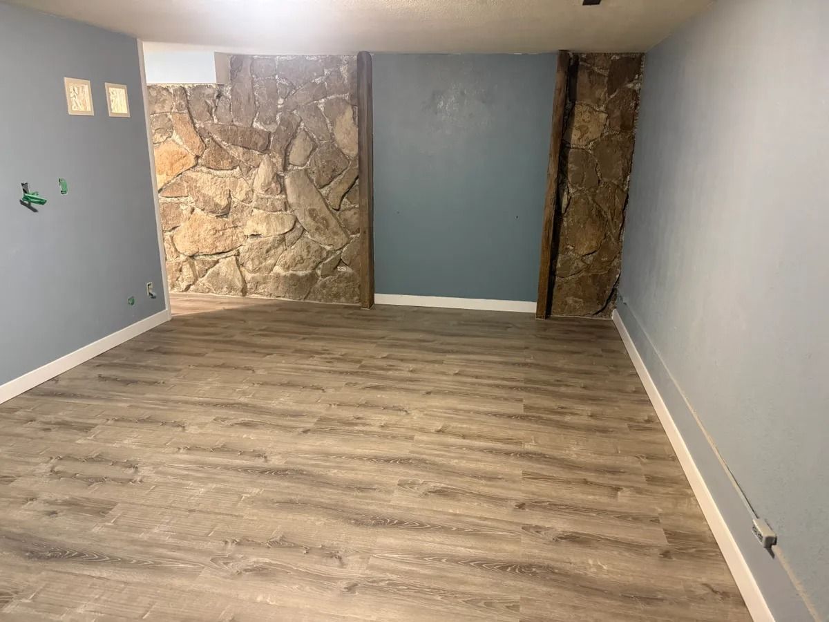 Empty basement room with wood floor, blue-gray walls, stone accent wall, and white ceiling lights