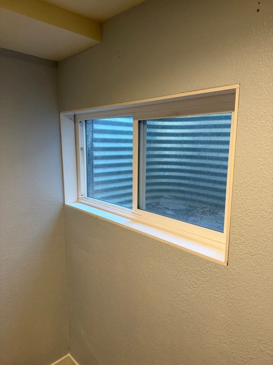 Small beige room with a rectangular window showing blue corrugated siding outside