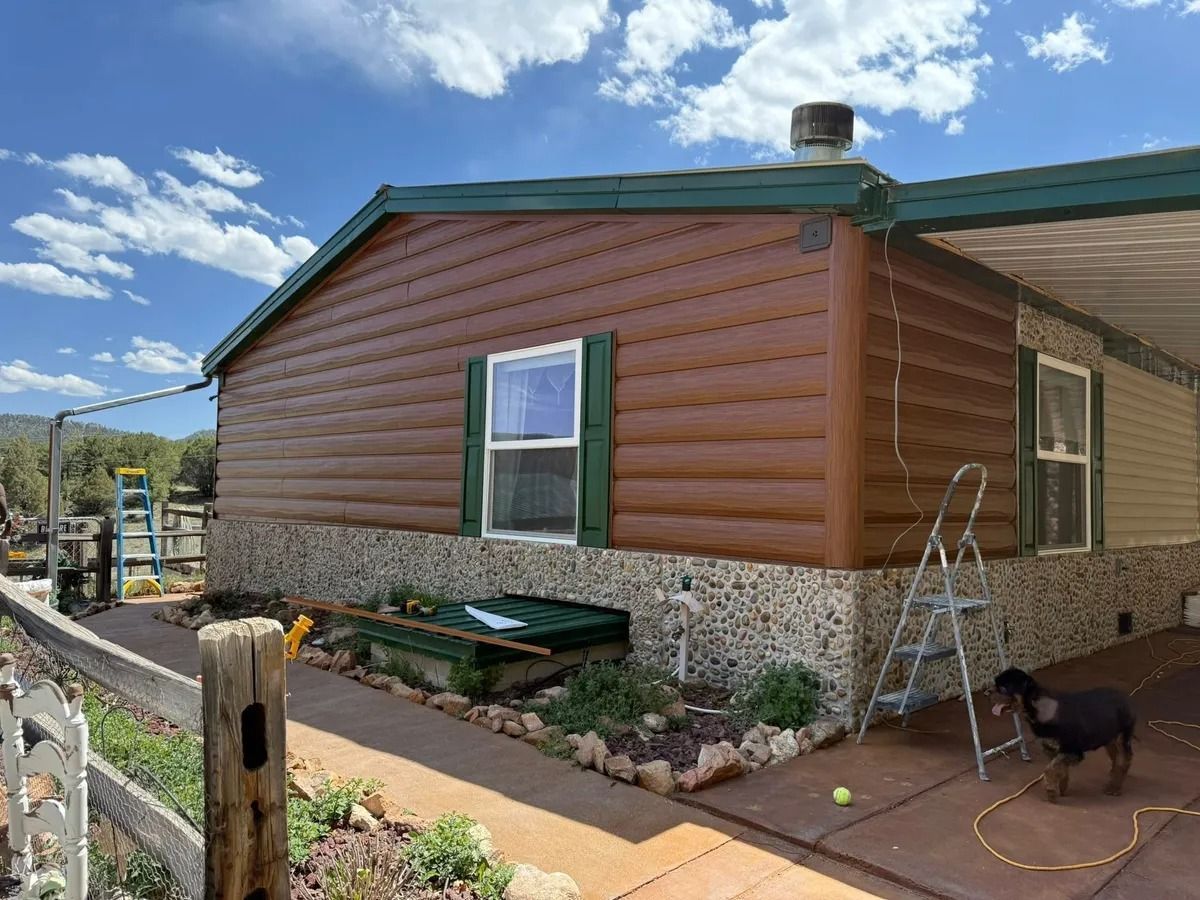 Mobile home with brown siding and stone base, ladder and tools outside, under a blue sky.