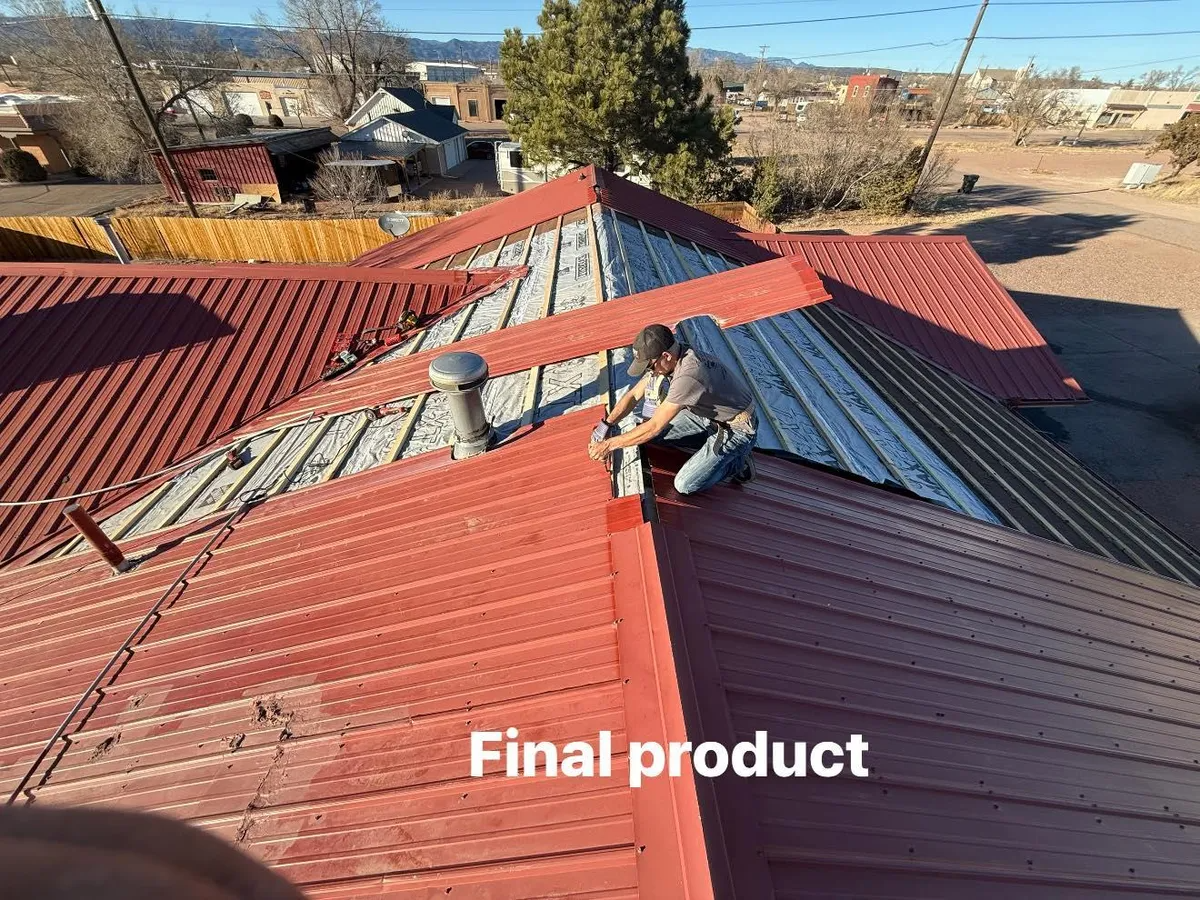 Two workers on a red metal roof, with one kneeling near a roof seam and distant desert houses in view