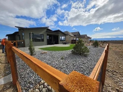 Modern house with gravel yard, small trees, and a wooden fence under a cloudy blue sky