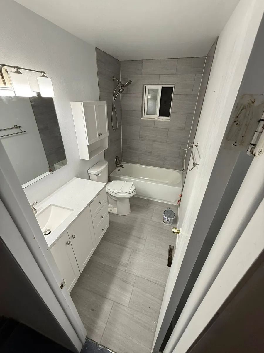 Small modern bathroom with gray tile, white vanity, toilet, tub, and mirrored cabinet.