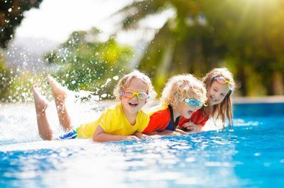 Three children in goggles splash and smile in a bright swimming pool outdoors.