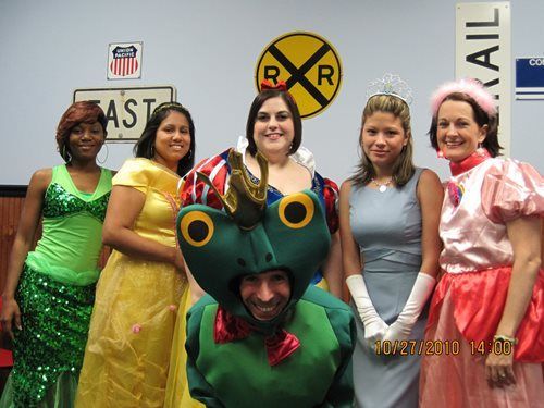 Five people pose indoors, including one in a frog costume and others in colorful outfits.