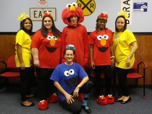Group of people in colorful Elmo and Cookie Monster costumes posing indoors with Sesame Street signs behind them