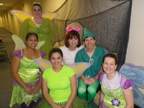 Five people in bright green and purple costumes pose indoors with fairy wings.