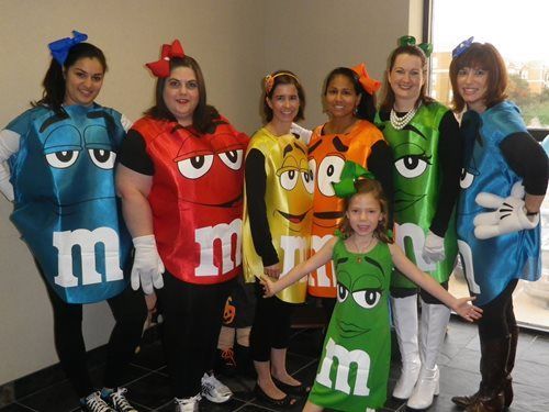 People in colorful M&M costumes pose together indoors, smiling around a child in a green M&M outfit
