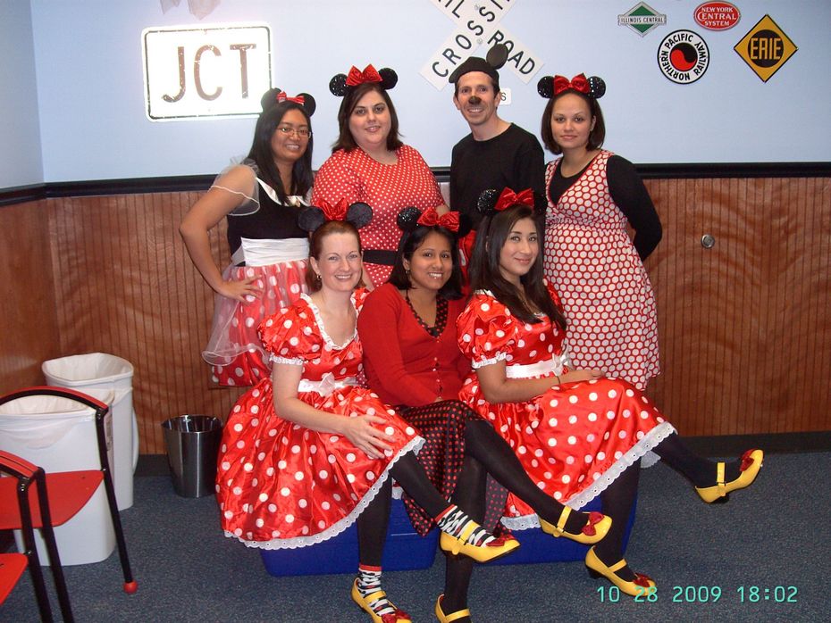 Six people in red polka-dot costumes posing indoors, four seated and two standing, with a dance club backdrop