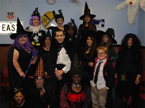 Group of people in Halloween costumes posing indoors, including witches, a devil, and a scarecrow.