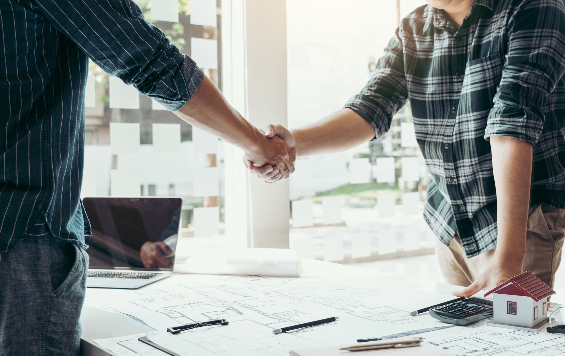 Two people shaking hands over architectural plans at a desk in a bright office