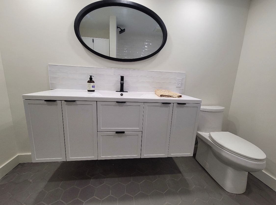 Modern bathroom with white vanity, oval mirror, double sinks, and toilet beside a tiled shower.