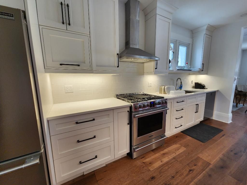 Bright white kitchen with stainless steel stove, hood, and wood floors, lit by under-cabinet lighting