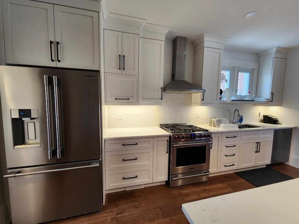 Modern white kitchen with stainless steel appliances, wood floor, and bright window over the sink.