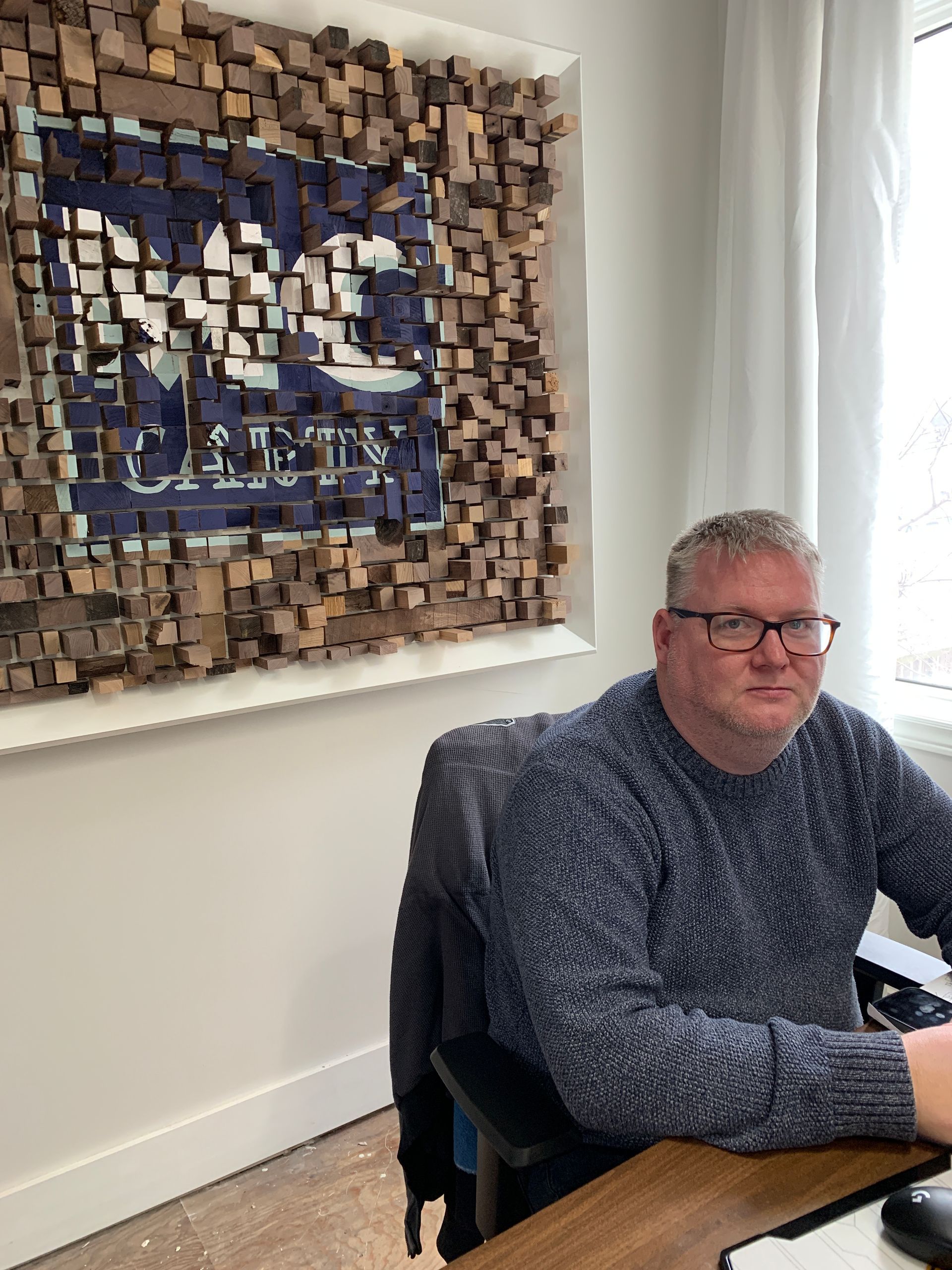 Man seated at a desk beside a textured wall artwork in a bright office.